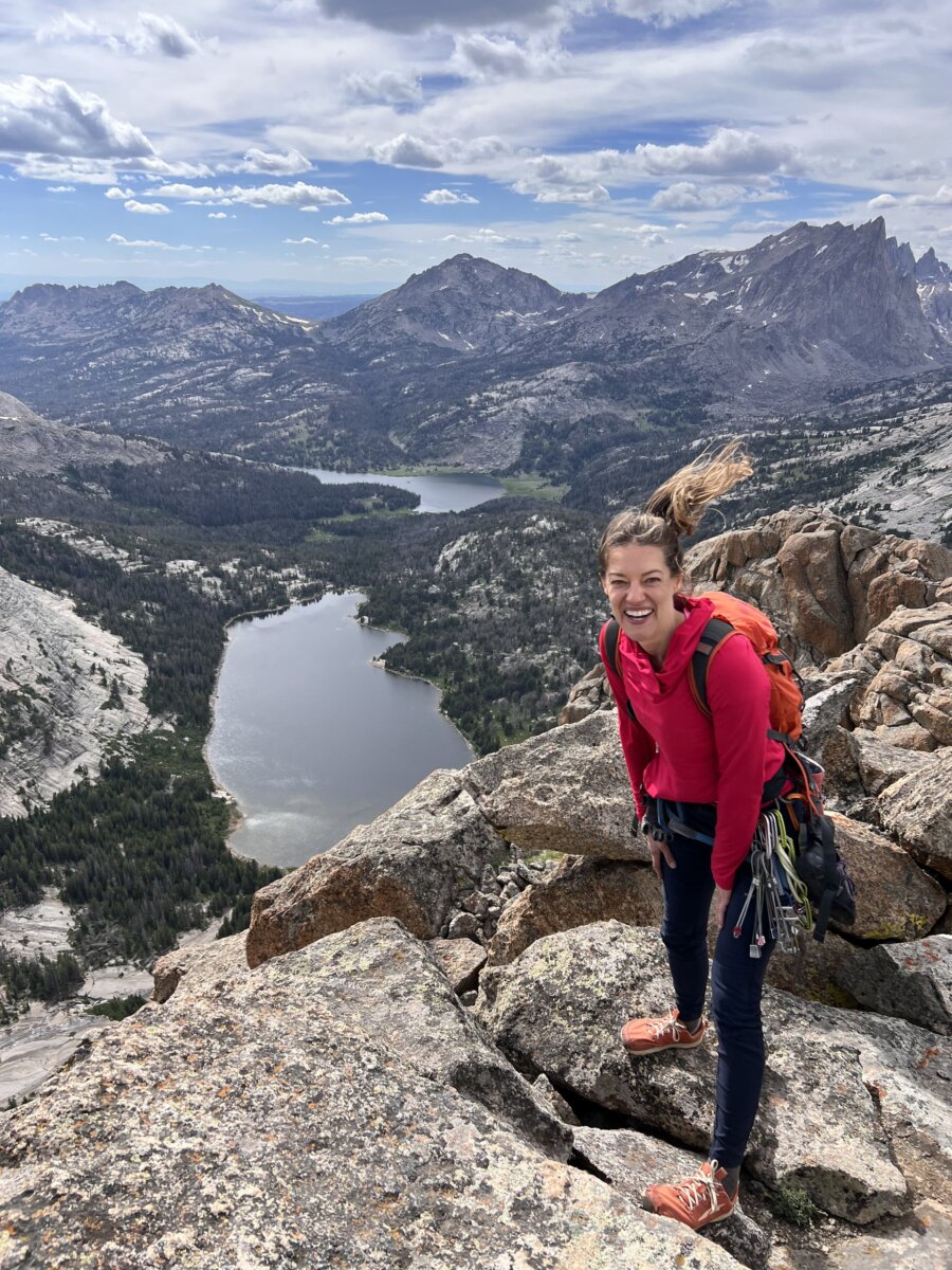 Laurel Graefe, dressed in hiking gear, stands on rocky terrain atop a mountain, smiling, with lakes and mountains visible in the background under a partly cloudy sky.