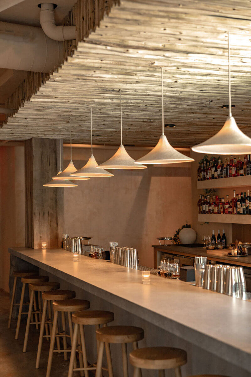 A modern bar with wooden stools, a concrete countertop, pendant lights, and shelves stocked with bottles and glassware—expertly curated by interior designer Alexandros Darsinos to showcase exceptional interior design.