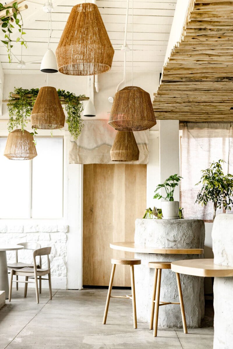 Modern café interior envisioned by Interior Designer Alexandros Darsinos, featuring woven pendant lights, wooden furniture, white walls, and potted plants. Natural light fills the space, creating a bright and airy atmosphere.