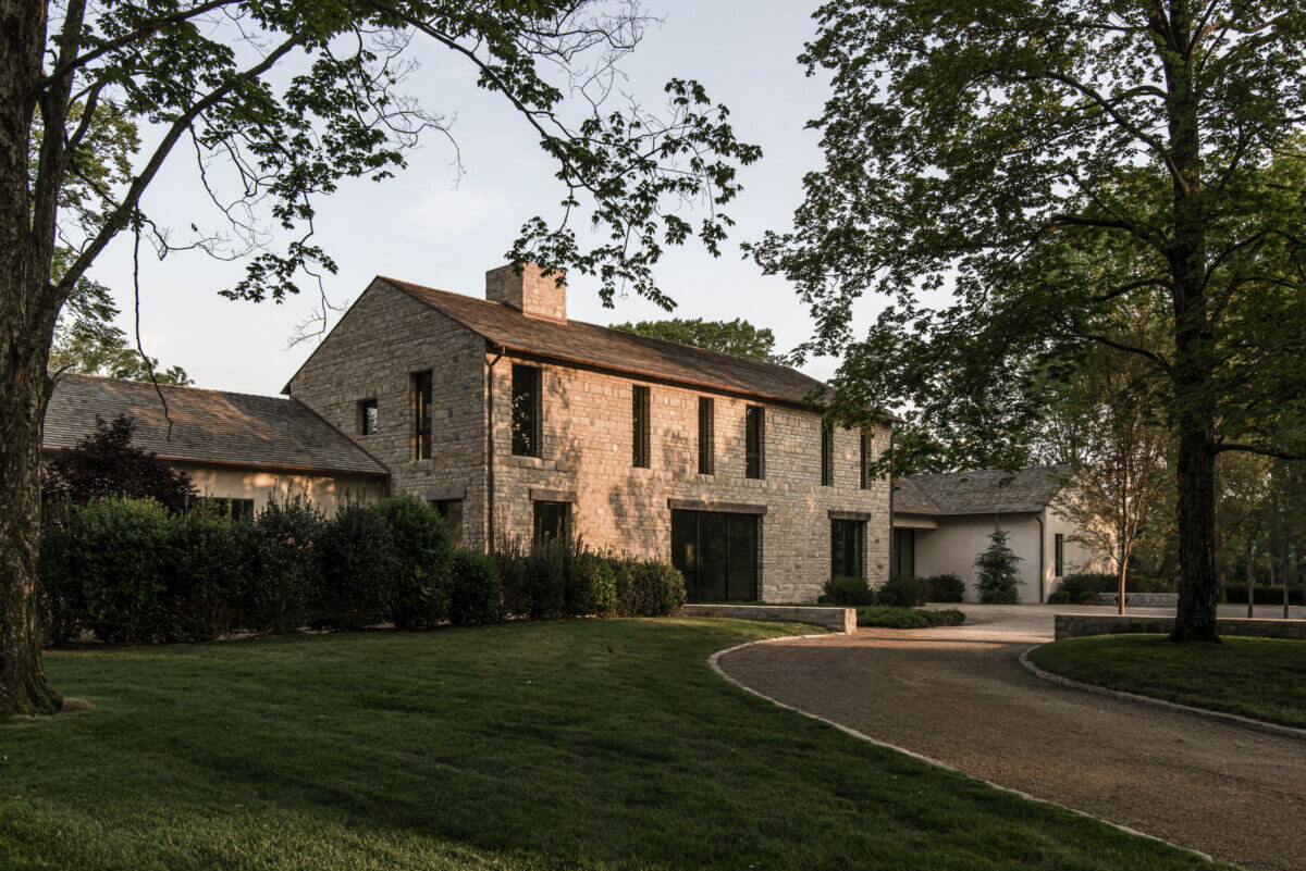 Two-story stone house with dark windows, a sloped roof, and a curved driveway, surrounded by trees and landscaped grass—an elegant residence envisioned to complement exquisite interior design curated by Alexandros Darsinos.
