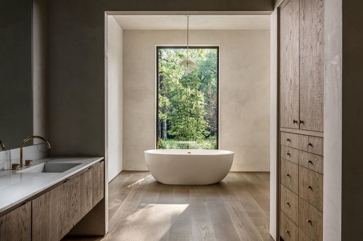 Modern bathroom with freestanding white bathtub, large window showing trees, wooden cabinetry, and a double sink with brass fixtures. Natural light fills the space, showcasing exquisite interior design by Alexandros Darsinos.