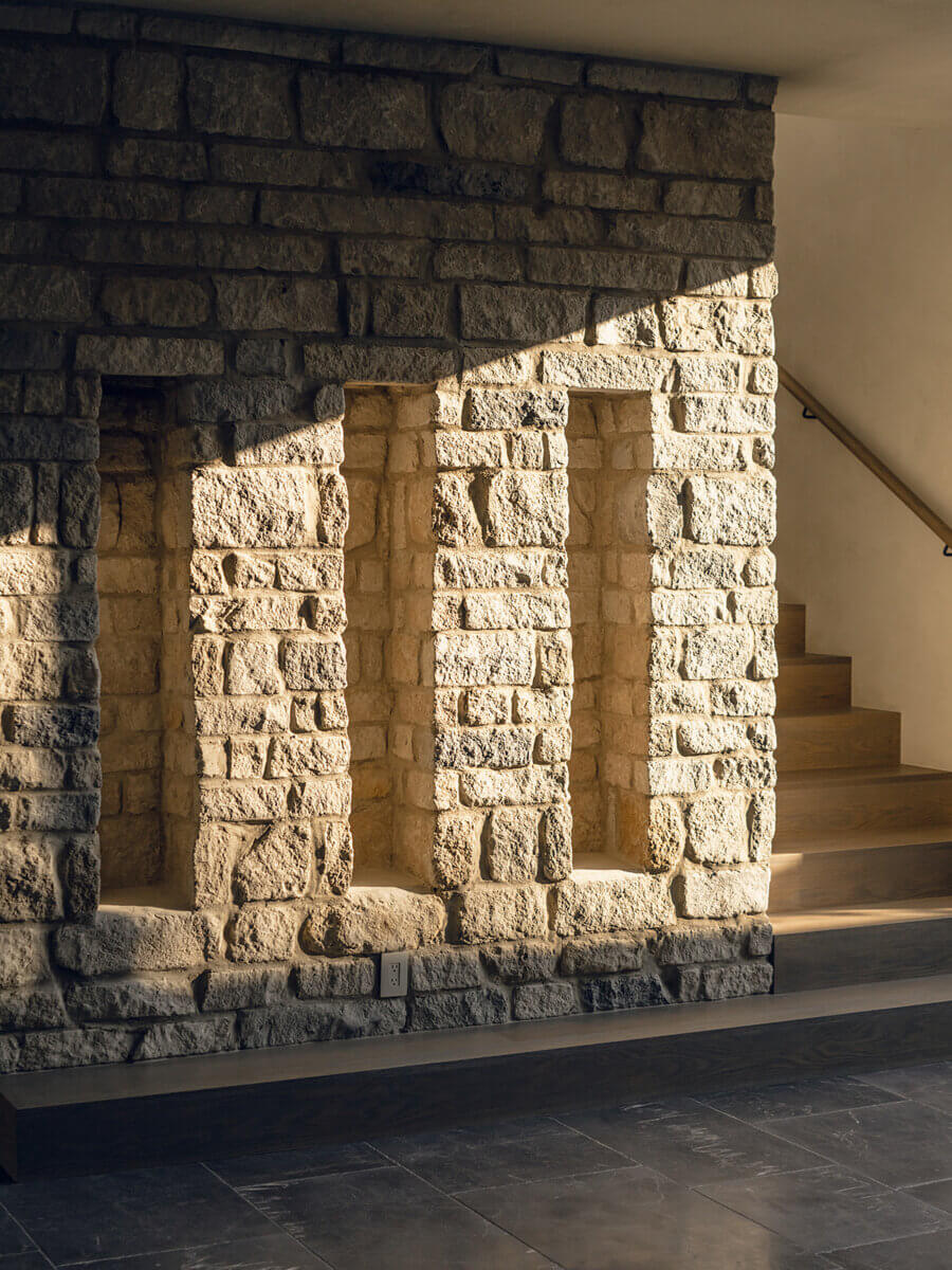 Sun shines on a textured stone wall with three rectangular niches, next to a wooden staircase—an elegant composition reflecting the expertise of Alexandros Darsinos, Interior Designer. The floor appears to be made of dark stone tiles, showcasing refined interior design.