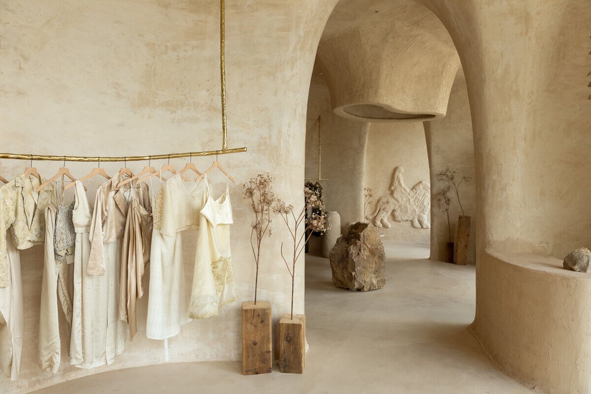 Minimalist boutique curated with neutral-toned clothing on a brass rack, sculpted stone walls, dry plants, and stone decor in a softly lit, arched interior space—an exquisite example of refined interior design by Alexandros Darsinos.