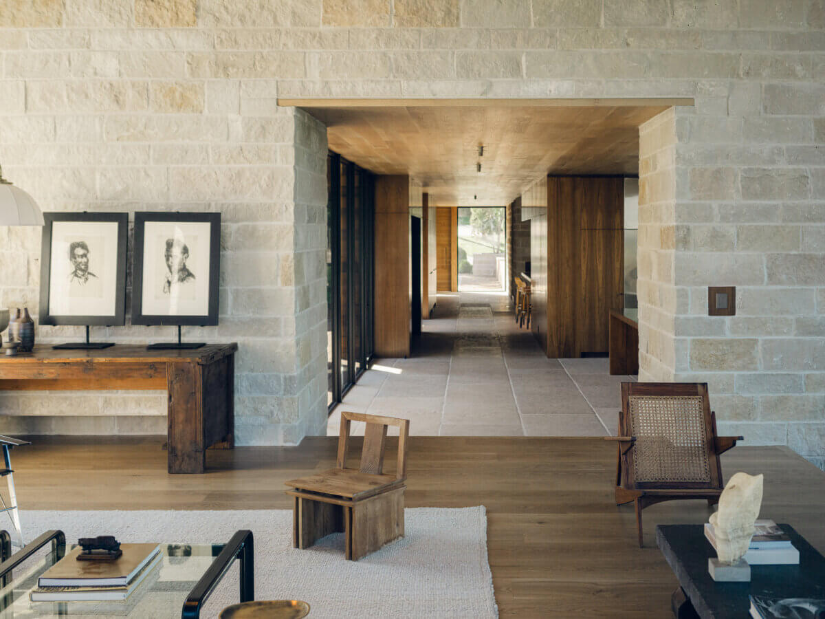 Modern living space with stone walls, wooden furniture, framed sketches on a console, and a hallway leading to another room. Thoughtfully curated by interior designer Alexandros Darsinos, the room’s natural light enters from large windows on the left, showcasing inspired interior design elements throughout.