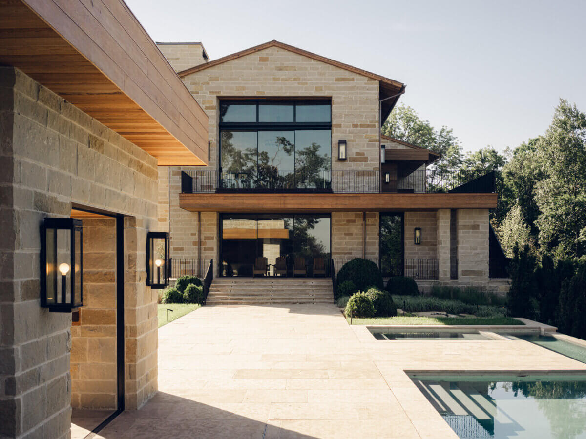 Modern stone house with large windows, wooden accents, and a swimming pool in the foreground, expertly crafted by interior designer Alexandros Darsinos. Surrounded by landscaped greenery under a clear sky, this home showcases thoughtful interior design that seamlessly blends indoor comfort with natural beauty.