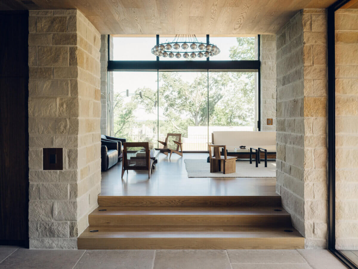 A modern living room with large windows, light stone walls, wooden floors, contemporary furniture, and a circular chandelier showcases the refined touch of interior designer Alexandros Darsinos. This inviting space exemplifies innovative interior design through its elegant materials and harmonious composition.