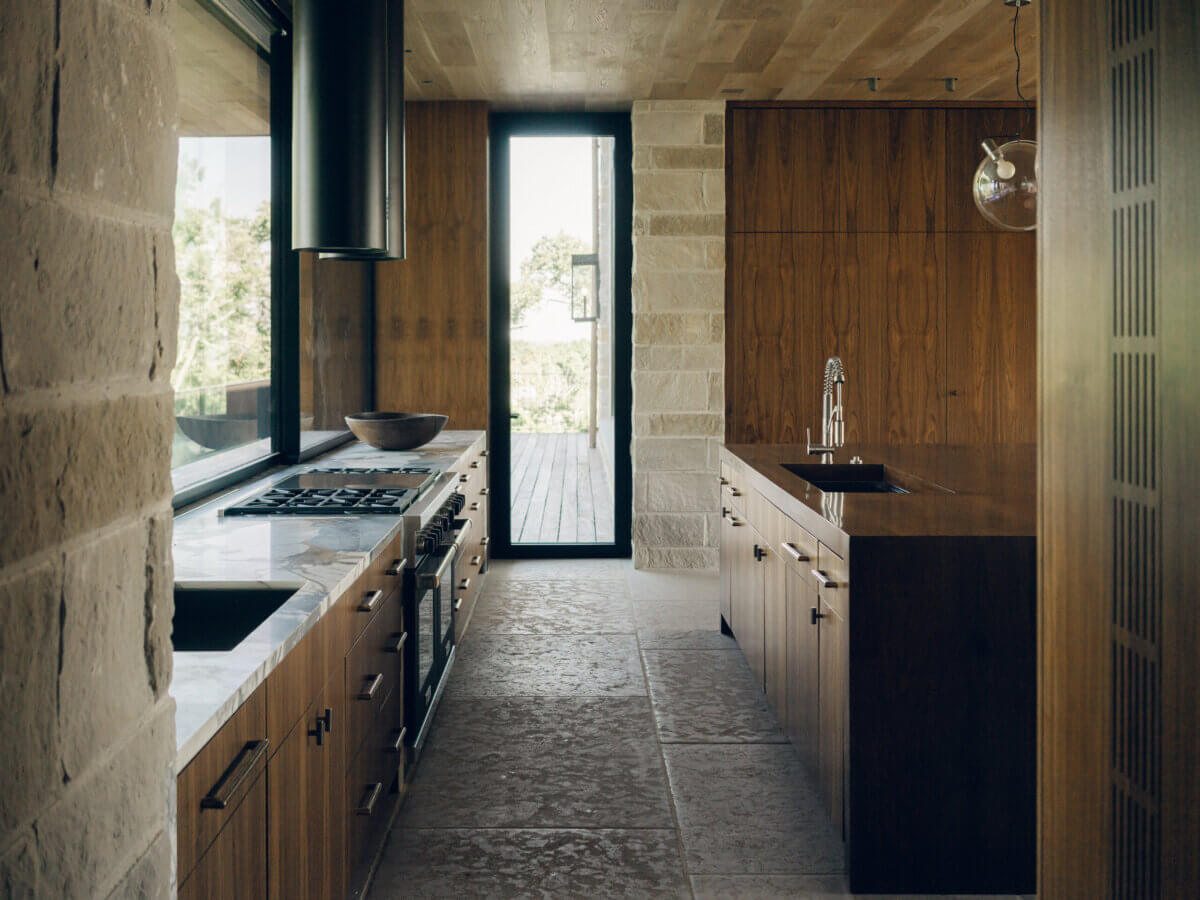 Modern kitchen with stone floors, wood cabinetry, marble countertops, stainless steel appliances, large window, and a glass door leading outside—expertly curated by Interior Designer Alexandros Darsinos to blend timeless elegance with functional design.