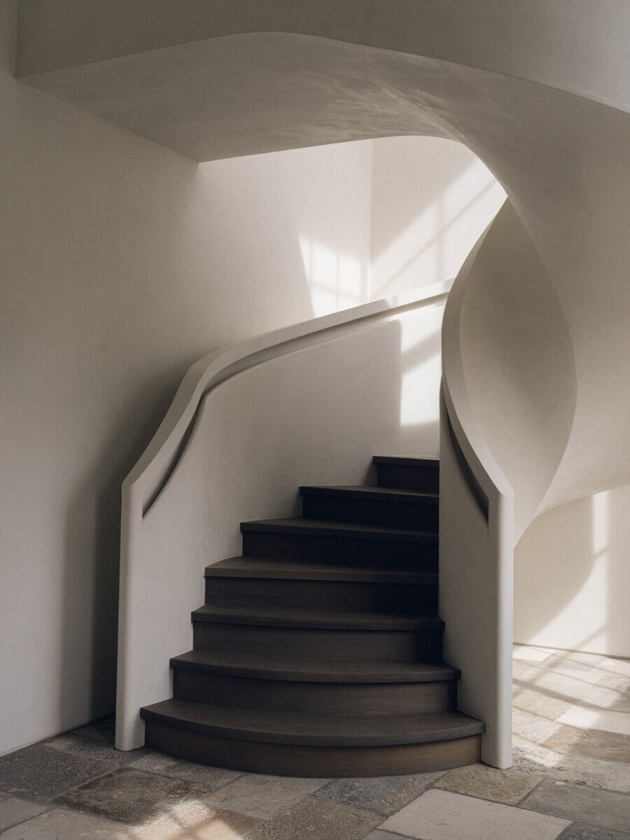 Curved wooden staircase with white plaster walls, illuminated by natural light from a nearby window, on a stone tile floor—a refined space expertly envisioned by interior designer Alexandros Darsinos.