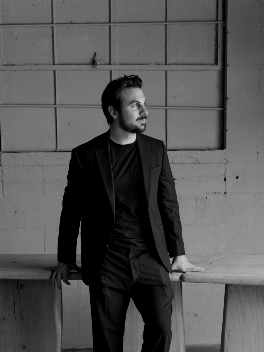 A man in a black suit stands indoors, leaning against a wooden table, looking to the side. The background features a concrete wall and a large, square-paned window—an elegant setting that reflects the refined eye of an interior designer. Black and white photo.