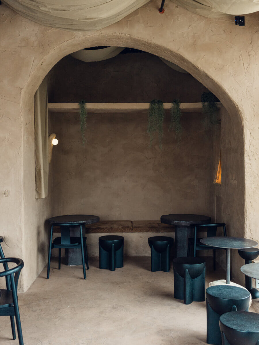 Minimalist cafe interior envisioned by Interior Designer Alexandros Darsinos, featuring an arched doorway, muted beige walls, dark modern chairs and tables, a built-in bench, and hanging greenery accents.