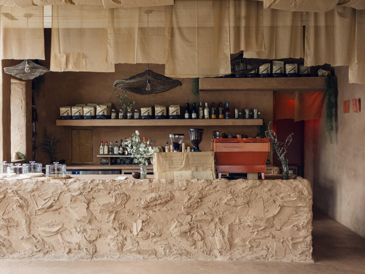 A rustic café counter with a textured clay front, shelves holding coffee bags and bottles, a red espresso machine, and neutral-toned hanging fabrics overhead showcases the signature style of Interior Designer Alexandros Darsinos.