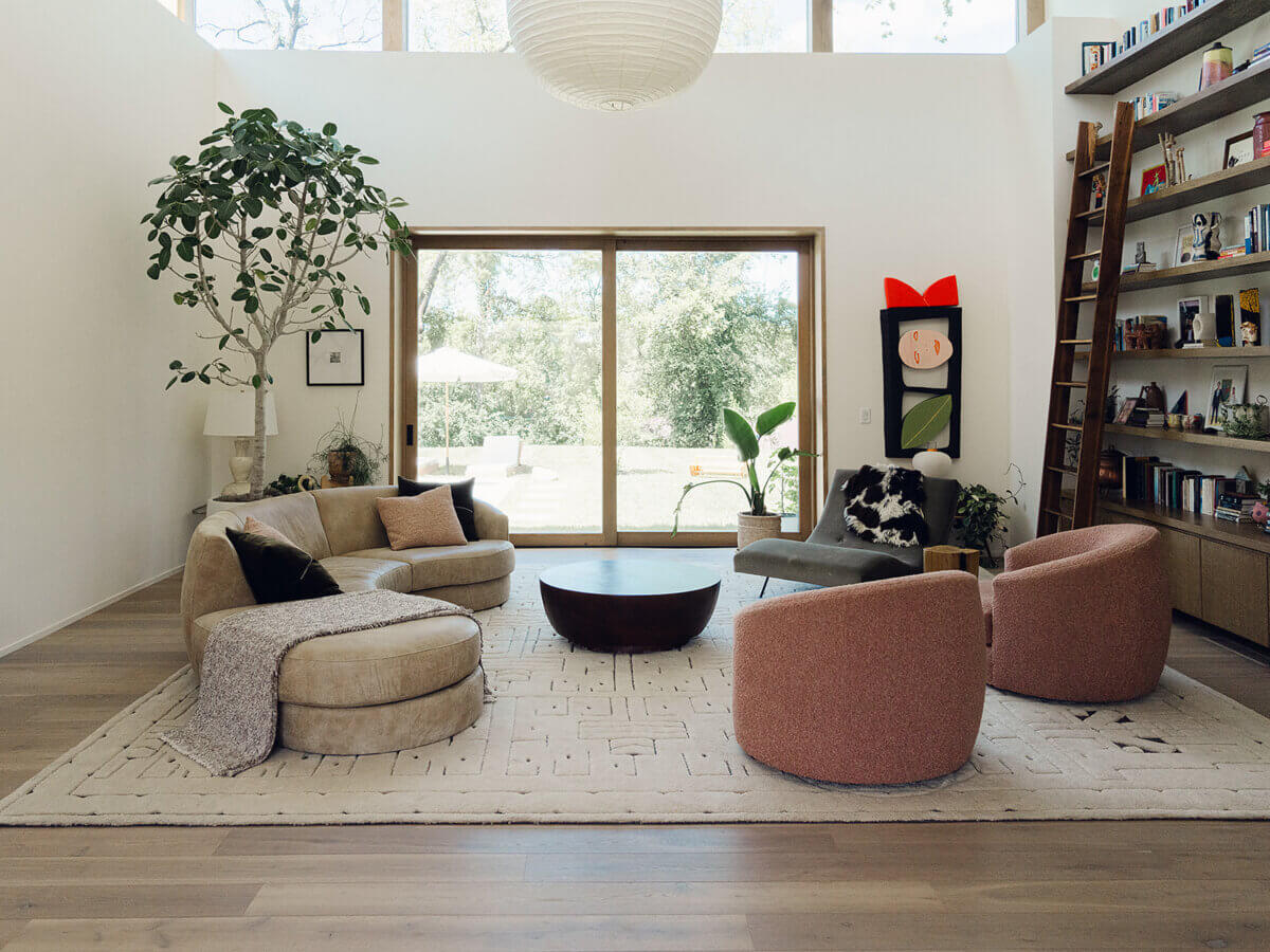 Modern living room with beige and pink chairs, a round coffee table, large rug, tall plant, wall art, bookshelves with ladder, and glass doors opening to a green outdoor area—an elegant space that showcases the refined touch of interior designer Alexandros Darsinos.
