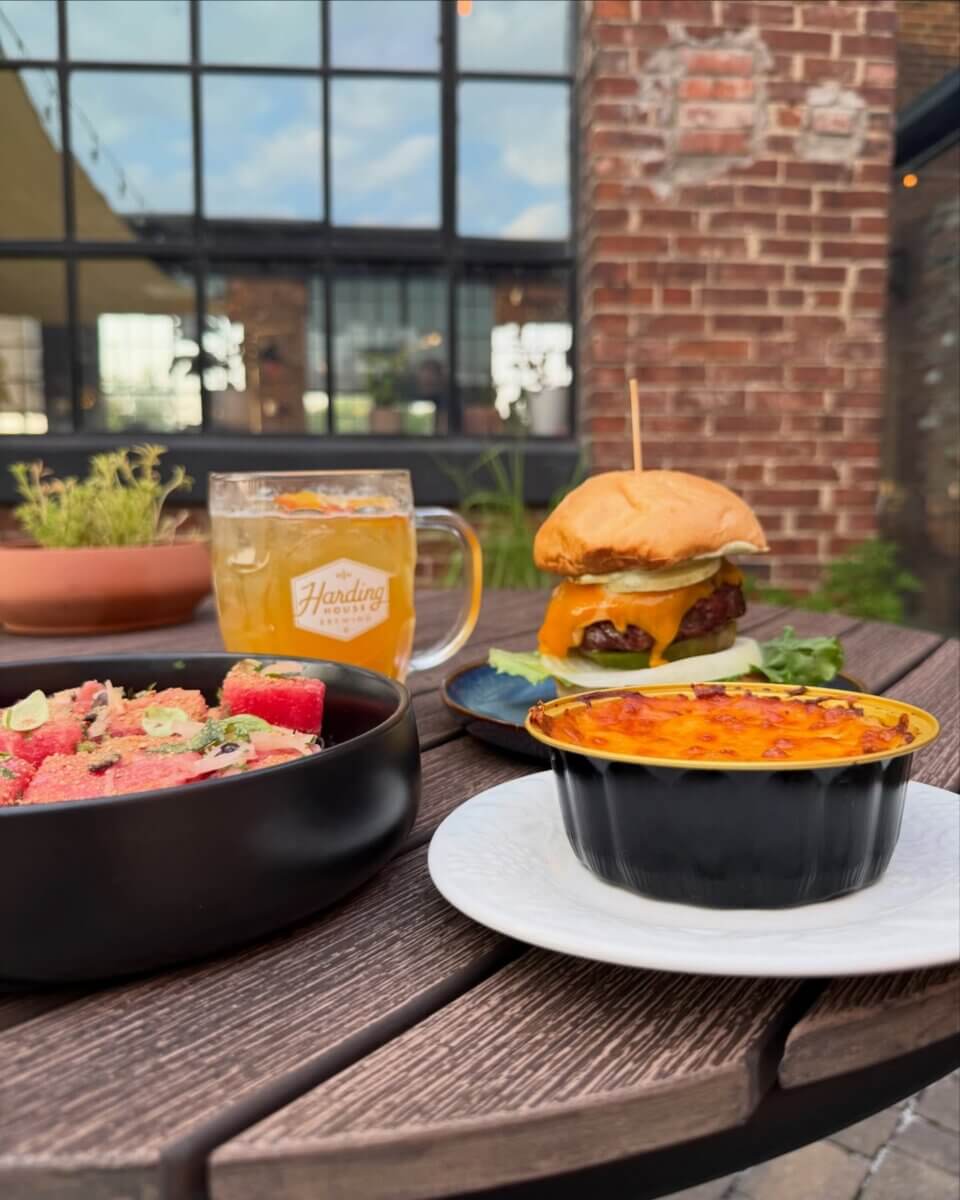 12 New Nashville Restaurants to Try This Month - 5 A wooden table with a watermelon salad, a burger with cheddar cheese, a baked dish, and a glass mug of beer, set outdoors near a brick building—capturing the laid-back charm of September evenings at Nashville restaurants.