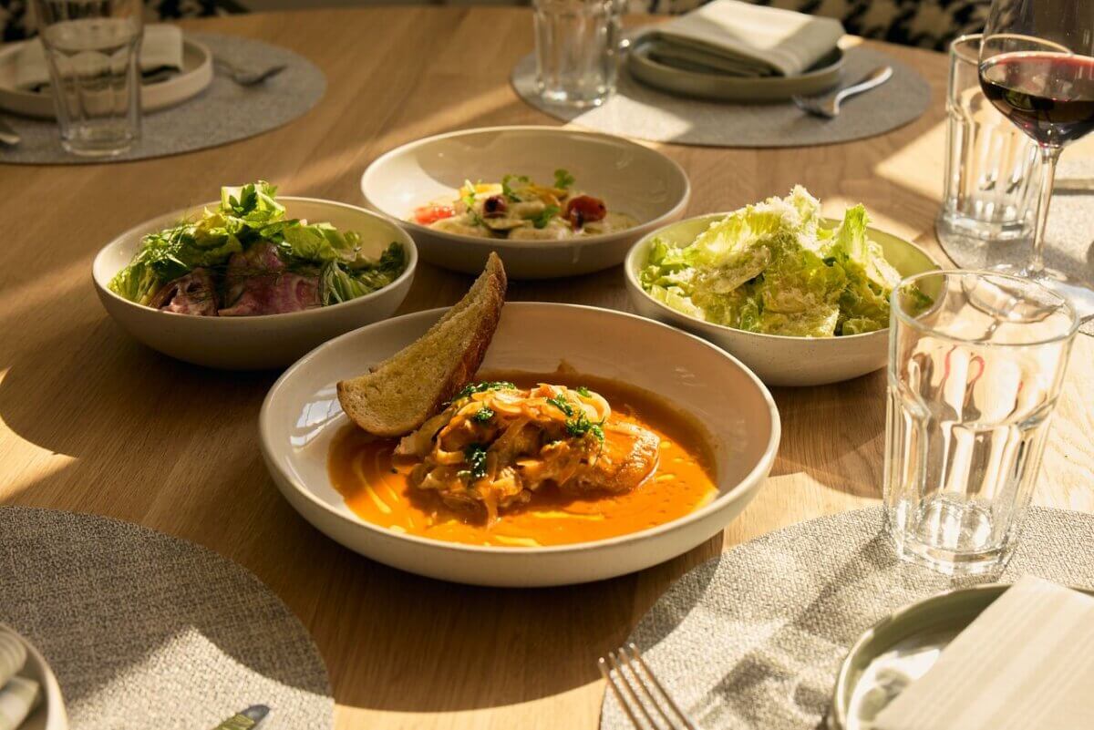 12 New Nashville Restaurants to Try This Month - 8 Four dishes on a wooden table: a bowl of soup with bread, two green salads, a tomato salad, empty plates, glasses, and a glass of red wine. Sunlight casts shadows over the scene—a picture-perfect September meal inspired by the vibrant offerings at some of the best new Nashville restaurants.