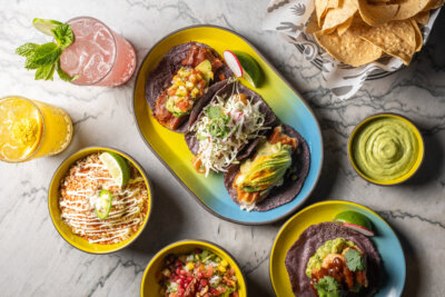 Three tacos on a blue and yellow plate, surrounded by bowls of elote, salsa, guacamole, chips, and two colorful drinks on a marble table—a vibrant spread you might find at new restaurants opening this September among standout Nashville restaurants.