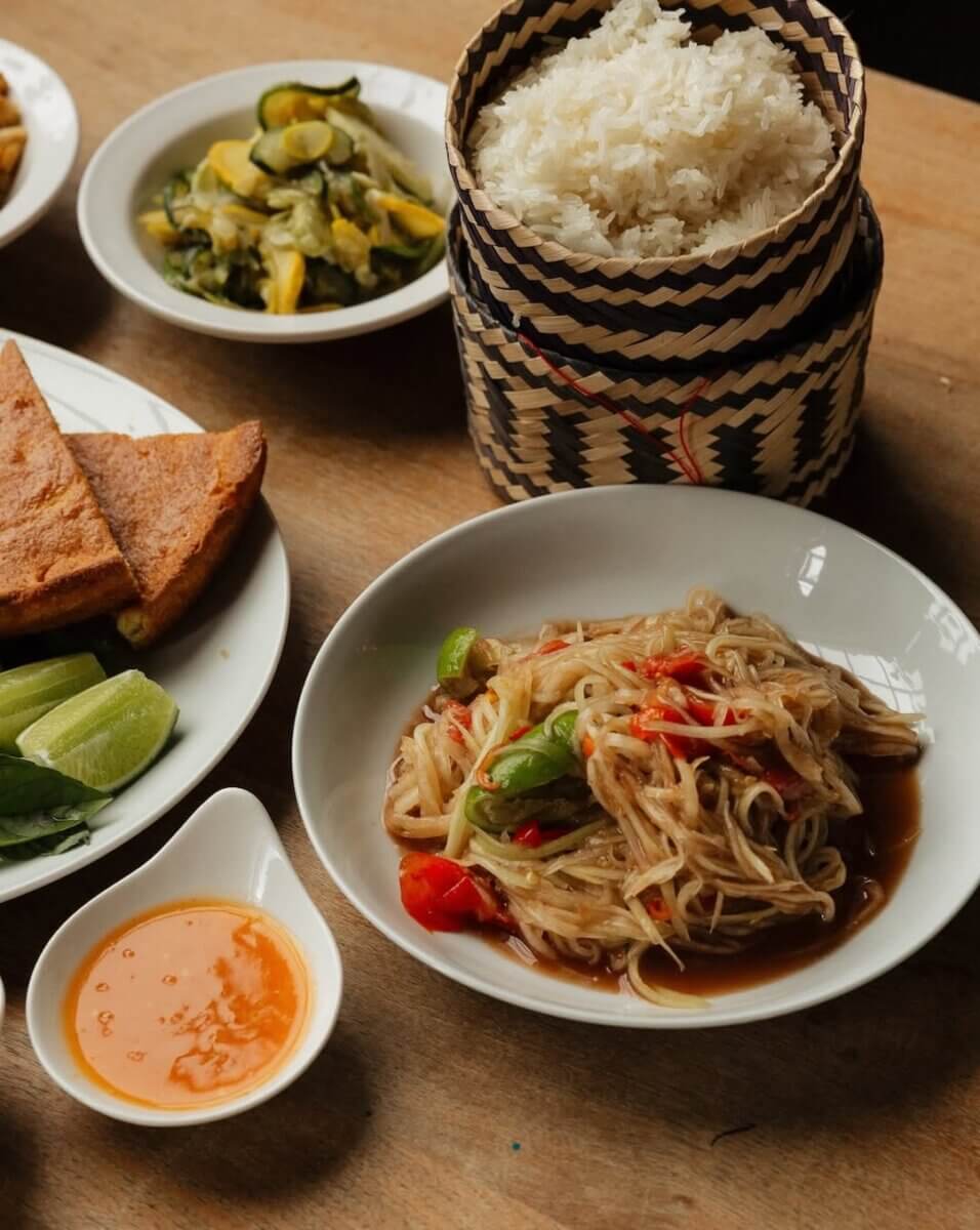 12 New Nashville Restaurants to Try This Month - 6 A plate of noodle salad with vegetables, sticky rice in a woven container, dipping sauce, and various side dishes on a wooden table—perfect for exploring the bold flavors offered by new restaurants opening in Nashville this September.