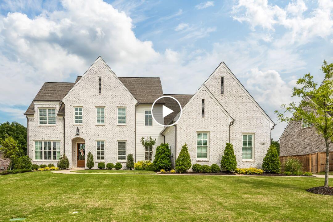 Large white brick house with steep gable roofs sits on a green lawn with shrubs, under a partly cloudy sky overhead. Located at 1548 Paso Fino Trail in Collierville, TN, this charming residence offers timeless curb appeal and a welcoming atmosphere.
