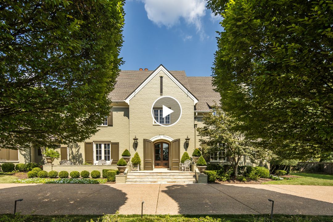 Two-story beige house with arched entrance, mansard roof, and symmetric landscaping, beautifully situated at 9320 Gwynn Hollow Cv, Germantown TN. Framed by tall trimmed trees under a sunny sky with some clouds.