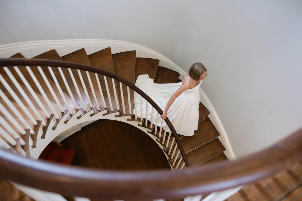 A Nashville Bride's Beautiful Tribute to Her Late Dad Only Made the Wedding More Joyful - 7 A woman in a white dress walks down a curved wooden staircase with white railings, seen from above, evoking the timeless elegance of a Brentwood Garden-Party Wedding at Ravenswood Mansion.