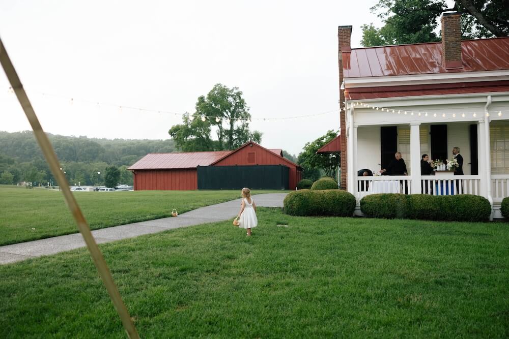 A Nashville Bride's Beautiful Tribute to Her Late Dad Only Made the Wedding More Joyful - 5 A young girl in a white dress walks across a grassy yard toward a red barn, with people gathered on a porch in the background, capturing the charming atmosphere of a garden-party wedding at Ravenswood Mansion in Brentwood.