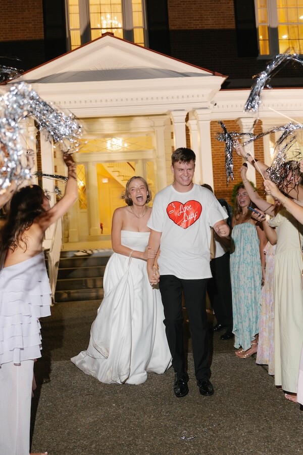 A Nashville Bride's Beautiful Tribute to Her Late Dad Only Made the Wedding More Joyful - 19 A bride and groom hold hands and smile while walking outside at their charming garden-party wedding in Brentwood, surrounded by guests waving pom-poms in celebration at the picturesque Ravenswood Mansion.