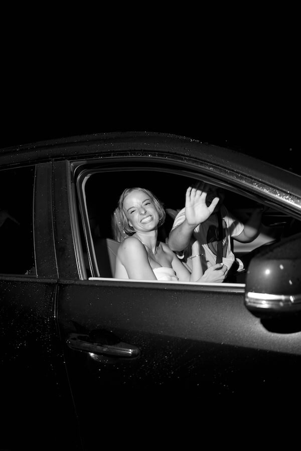 A Nashville Bride's Beautiful Tribute to Her Late Dad Only Made the Wedding More Joyful - 20 A woman smiles and waves from the front seat of a car at night, while another person next to her raises an arm, partially hiding their face—capturing a candid moment after a joyful Brentwood garden-party wedding.