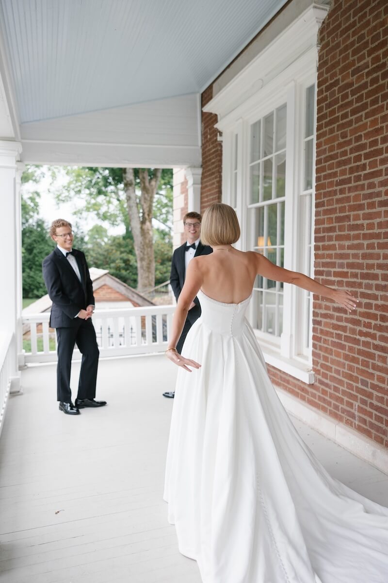 A Nashville Bride's Beautiful Tribute to Her Late Dad Only Made the Wedding More Joyful - 9 A bride in a white gown stands with arms open, facing two men in tuxedos on a porch with white railings and a brick wall, setting the scene for an elegant garden-party wedding at Ravenswood Mansion in Brentwood.