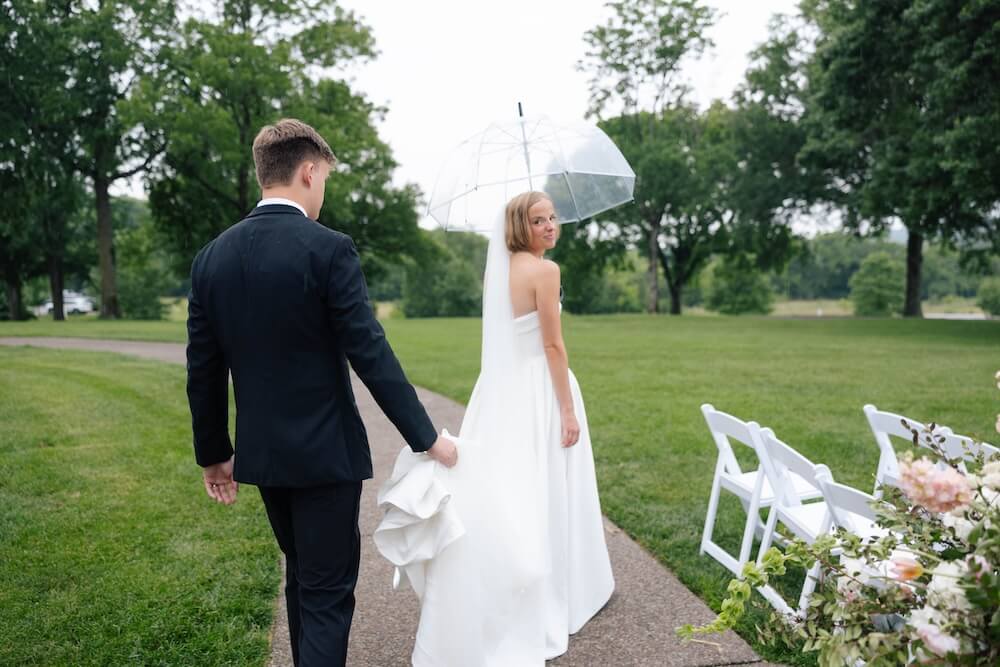A Nashville Bride's Beautiful Tribute to Her Late Dad Only Made the Wedding More Joyful - 4 A bride holding a clear umbrella walks on a path at Ravenswood Mansion while a groom in a suit follows, holding her dress. The setting is outdoors in Brentwood, surrounded by green grass, trees, and white chairs—perfect for a charming Garden-Party Wedding.