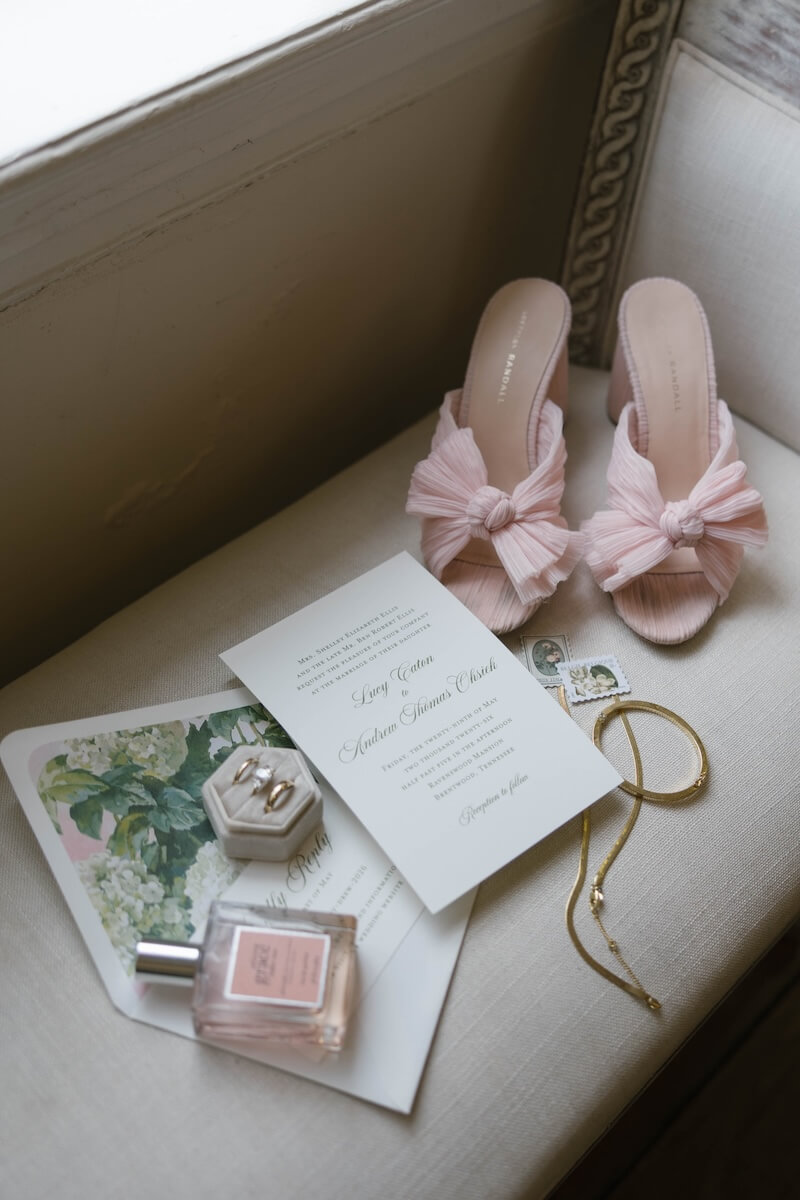 A Nashville Bride's Beautiful Tribute to Her Late Dad Only Made the Wedding More Joyful - 3 Pink heeled sandals, a wedding invitation, perfume, jewelry, vintage stamps, a floral envelope, and a ring box arranged on a cushioned bench evoke the timeless elegance of a garden-party wedding at Brentwood’s Ravenswood Mansion.