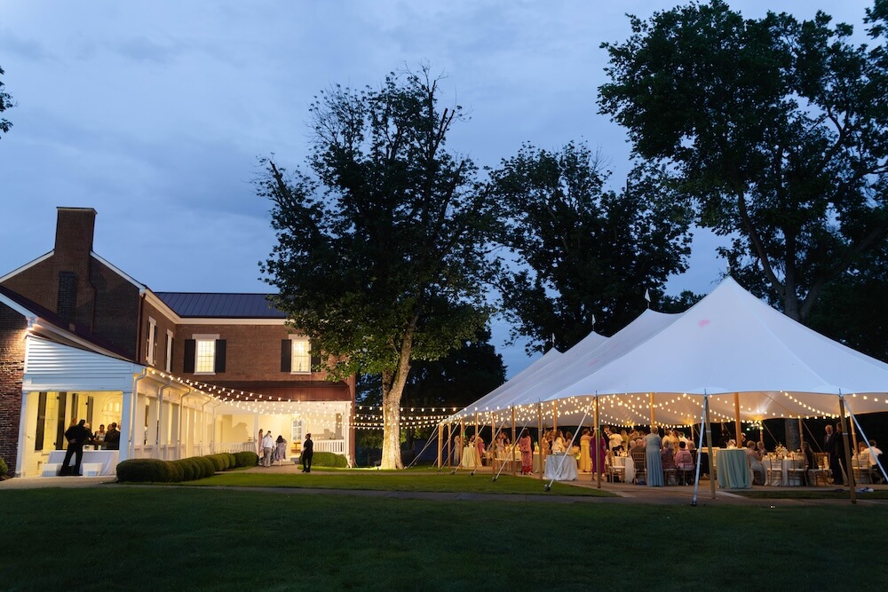 A Nashville Bride's Beautiful Tribute to Her Late Dad Only Made the Wedding More Joyful - 6 Large white event tent with string lights set up on a lawn beside a brick house at dusk, evoking the charm of a Garden-Party Wedding; guests are gathered inside and around the tent at Ravenswood Mansion in Brentwood.