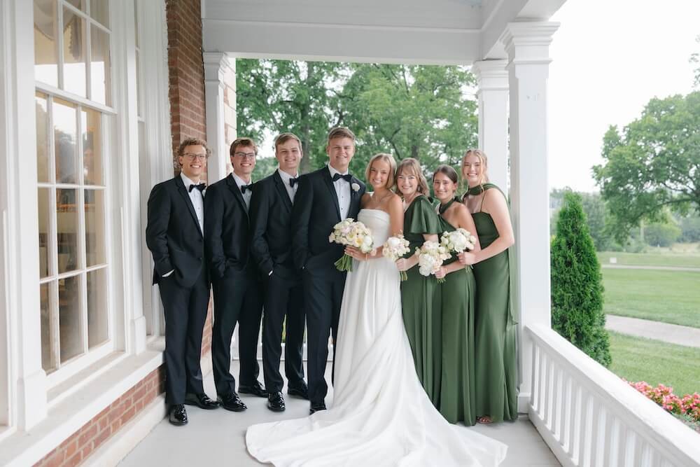 A Nashville Bride's Beautiful Tribute to Her Late Dad Only Made the Wedding More Joyful - 11 Six people in formal attire pose on a porch at Ravenswood Mansion in Brentwood; three men in black tuxedos and three women in green dresses join the bride, who stands out in a white gown holding bouquets—capturing the elegance of a garden-party wedding.