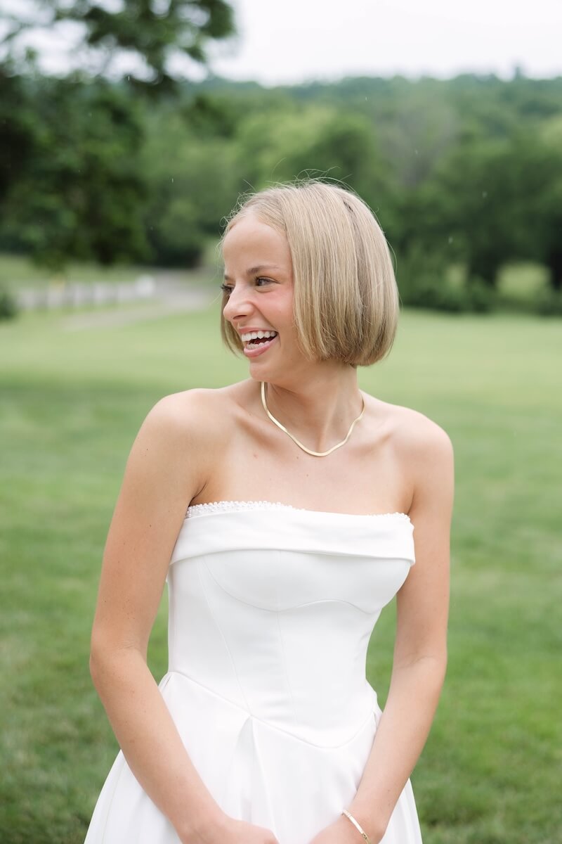 A Nashville Bride's Beautiful Tribute to Her Late Dad Only Made the Wedding More Joyful - 8 A young woman in a strapless white dress stands outdoors on a grassy lawn at Ravenswood Mansion, smiling and looking to the side, capturing the joyful spirit of a Brentwood garden-party wedding.