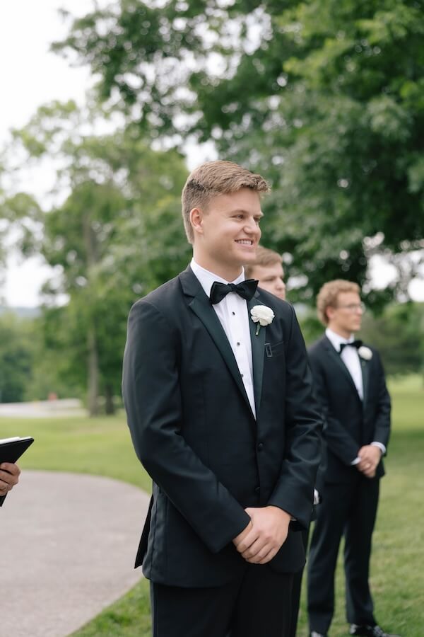A Nashville Bride's Beautiful Tribute to Her Late Dad Only Made the Wedding More Joyful - 14 A young man in a black tuxedo with a white boutonniere stands outdoors at a garden-party wedding, smiling, with two men in similar attire behind him and trees in the background at Ravenswood Mansion in Brentwood.