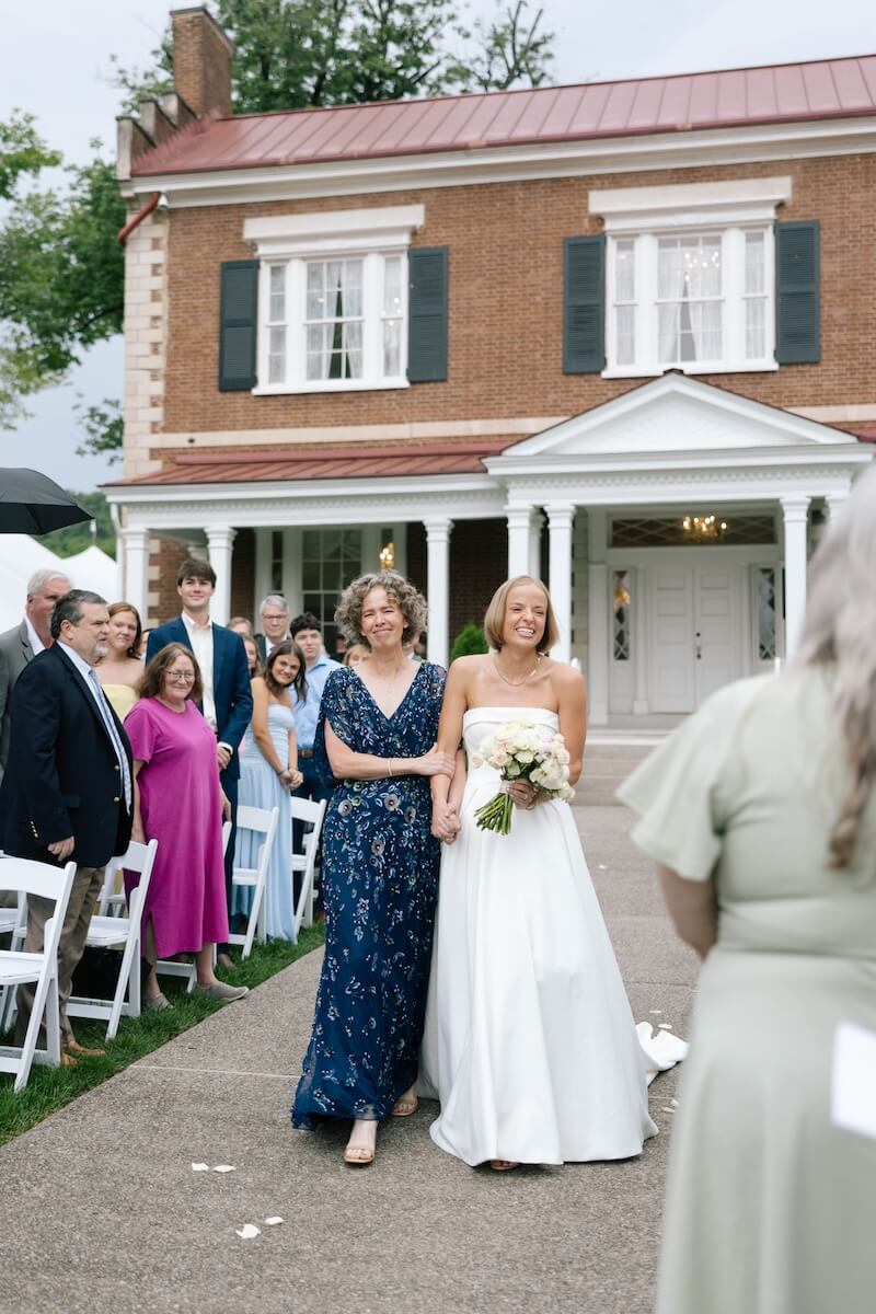 A Nashville Bride's Beautiful Tribute to Her Late Dad Only Made the Wedding More Joyful - 12 A bride in a white dress walks arm in arm with a woman in blue down an outdoor aisle, surrounded by seated guests in front of a brick building, capturing the charming ambiance of a garden-party wedding at Ravenswood Mansion in Brentwood.