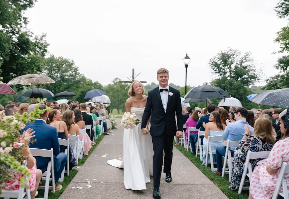 A Nashville Bride's Beautiful Tribute to Her Late Dad Only Made the Wedding More Joyful - 2 A bride and groom walk down the aisle outdoors at Ravenswood Mansion in Brentwood, smiling and holding hands during their enchanting garden-party wedding, while guests seated on both sides hold umbrellas.