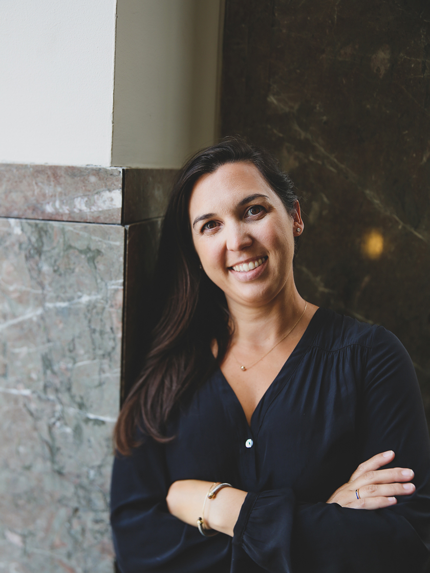 Woman with long dark hair, wearing a black blouse, stands with arms crossed and smiles while leaning against a marble wall indoors at 4000 Wallace Lane, Nashville.