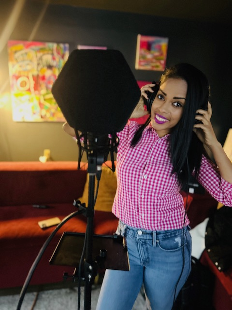 Woman artist in a pink checkered shirt and jeans stands at a microphone wearing headphones in a colorful, art-filled room, surrounded by vibrant visual arts.