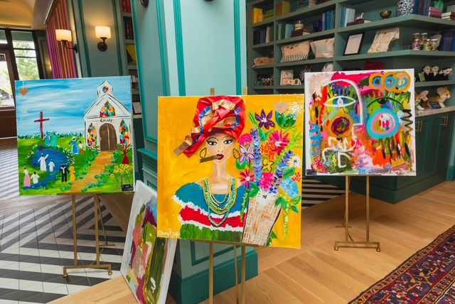Three colorful paintings on easels are displayed in a bright room with blue walls, patterned floors, and bookshelves in the background—a perfect setting for any art enthusiast or admirer of visual arts.