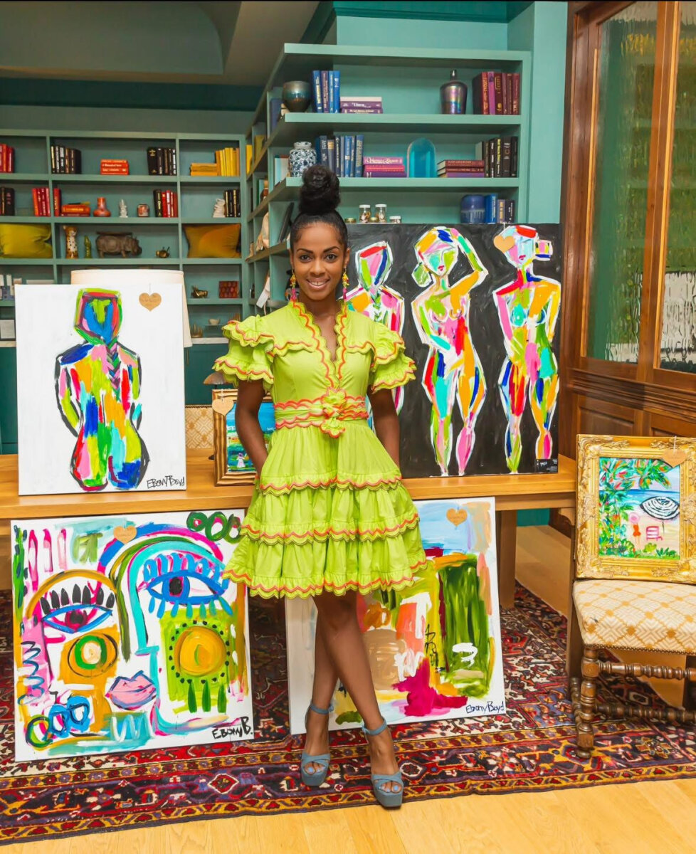 A woman artist in a bright green ruffled dress stands indoors, surrounded by colorful abstract paintings displayed on easels and a chair, with shelves of books and decor in the background—an inspiring scene that beautifully captures her passion for art appreciation.