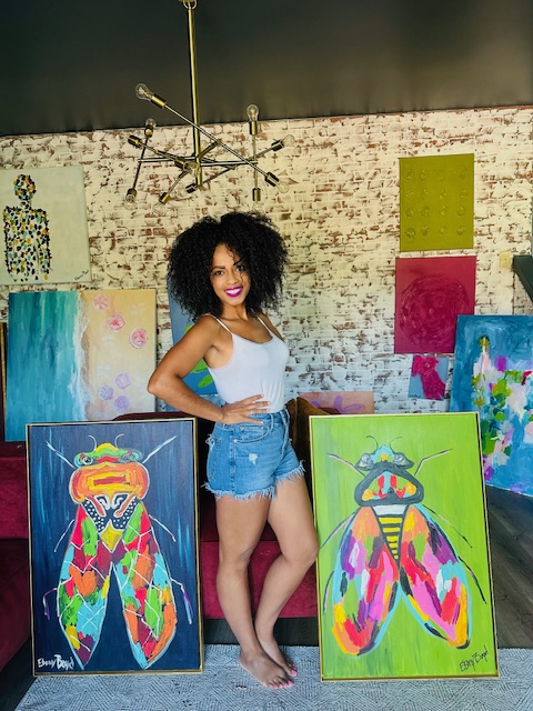 A woman artist in a white tank top and denim shorts stands indoors, smiling between two colorful insect paintings; various other artworks are displayed in the background, reflecting her passion for visual arts.