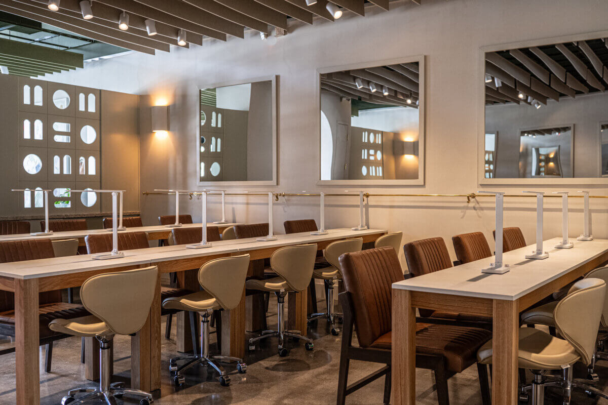 A modern nail salon with long tables, beige swivel chairs, desk dividers, and large mirrors on the walls under ceiling lights.
