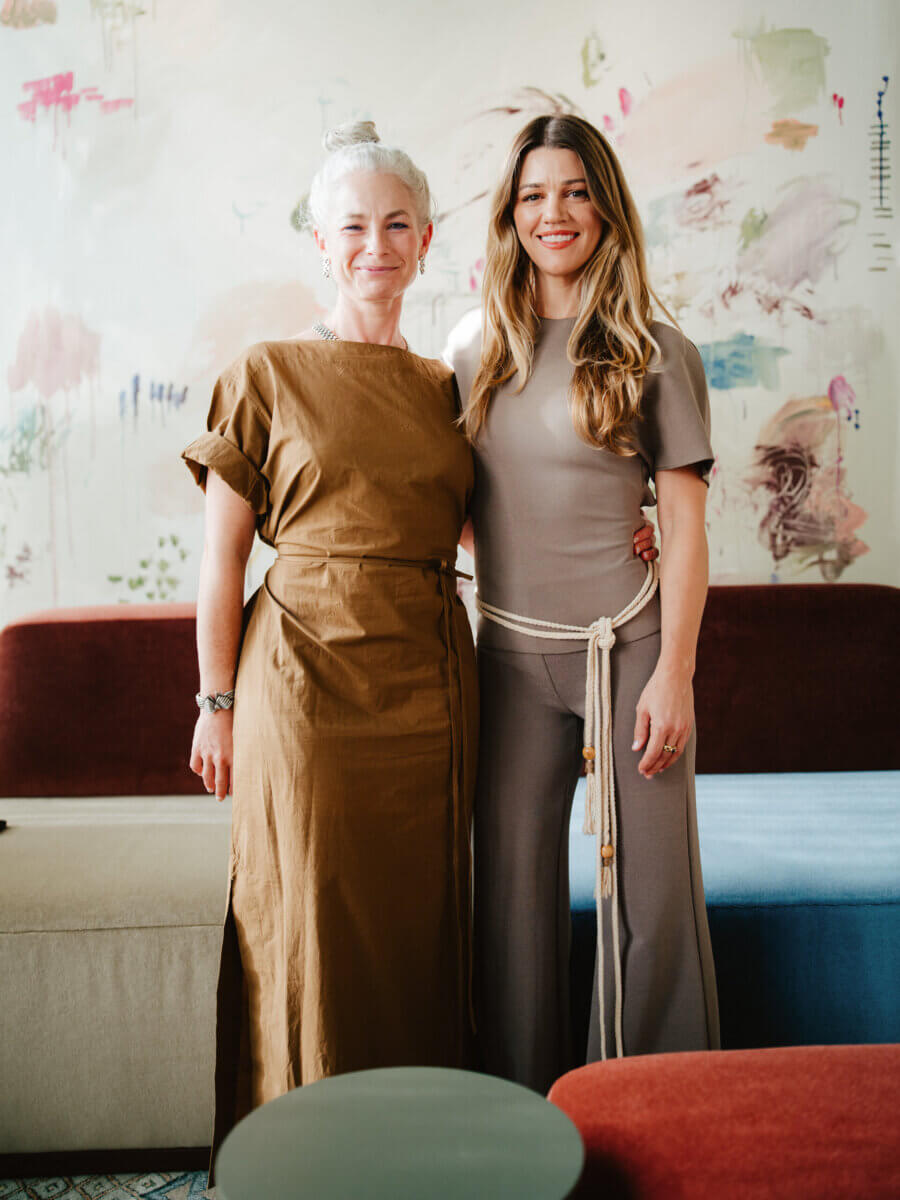 Two women stand side by side, smiling, in a modern room with abstract art on the wall behind them. One wears a brown dress, the other a taupe outfit with a rope belt.