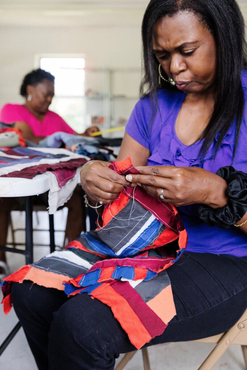 A woman in a purple shirt sews colorful fabric strips together, crafting a vibrant quilt—a scene that reflects the creative spirit found on many Alabama getaways—while another woman works in the background at a table.