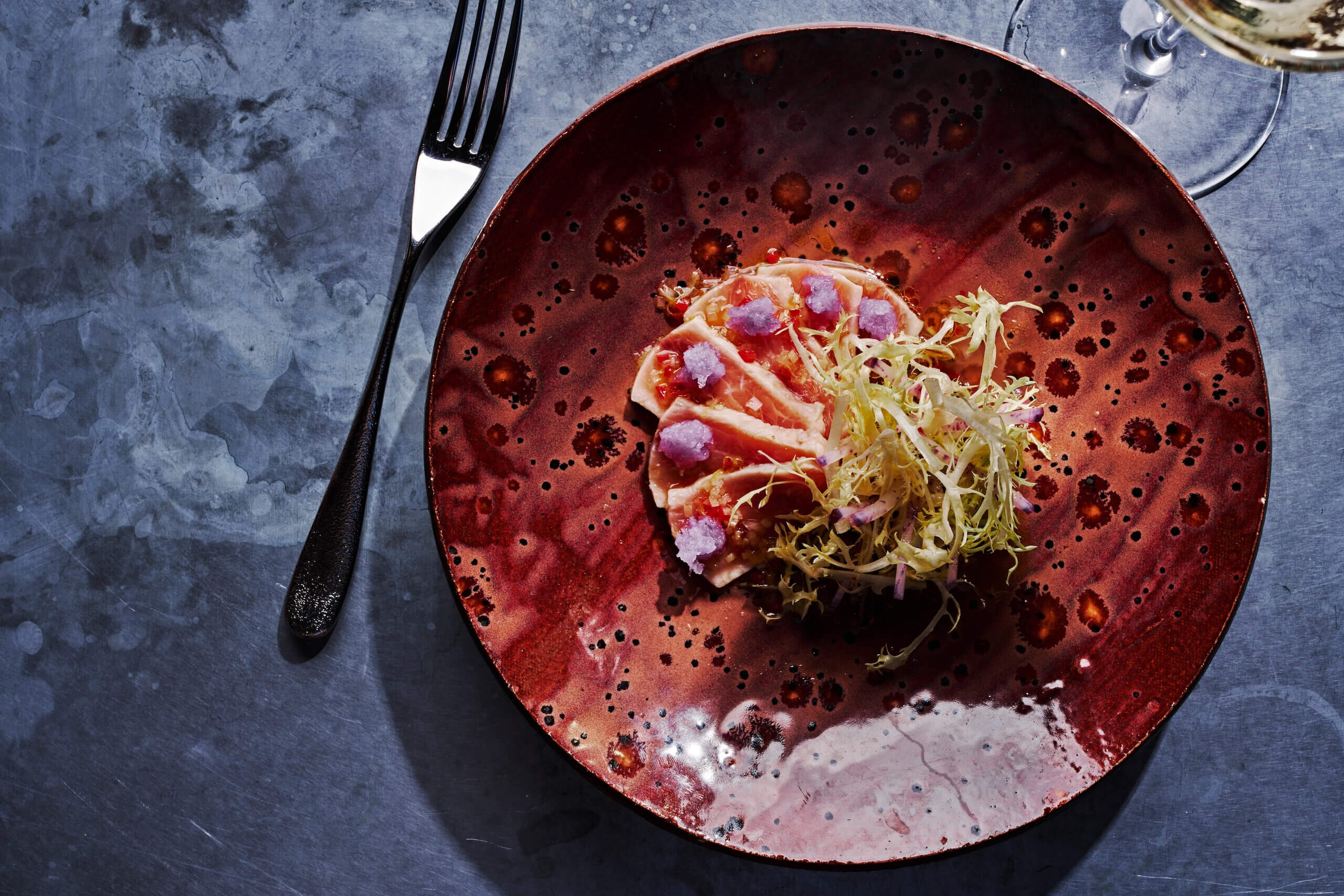 The Best Culinary Hot Spots in the South - 6 A red ceramic plate with a composed salad of greens, thinly sliced vegetables, and purple garnish, next to a fork and a glass—just the kind of fresh presentation you might find at the best southern restaurants.