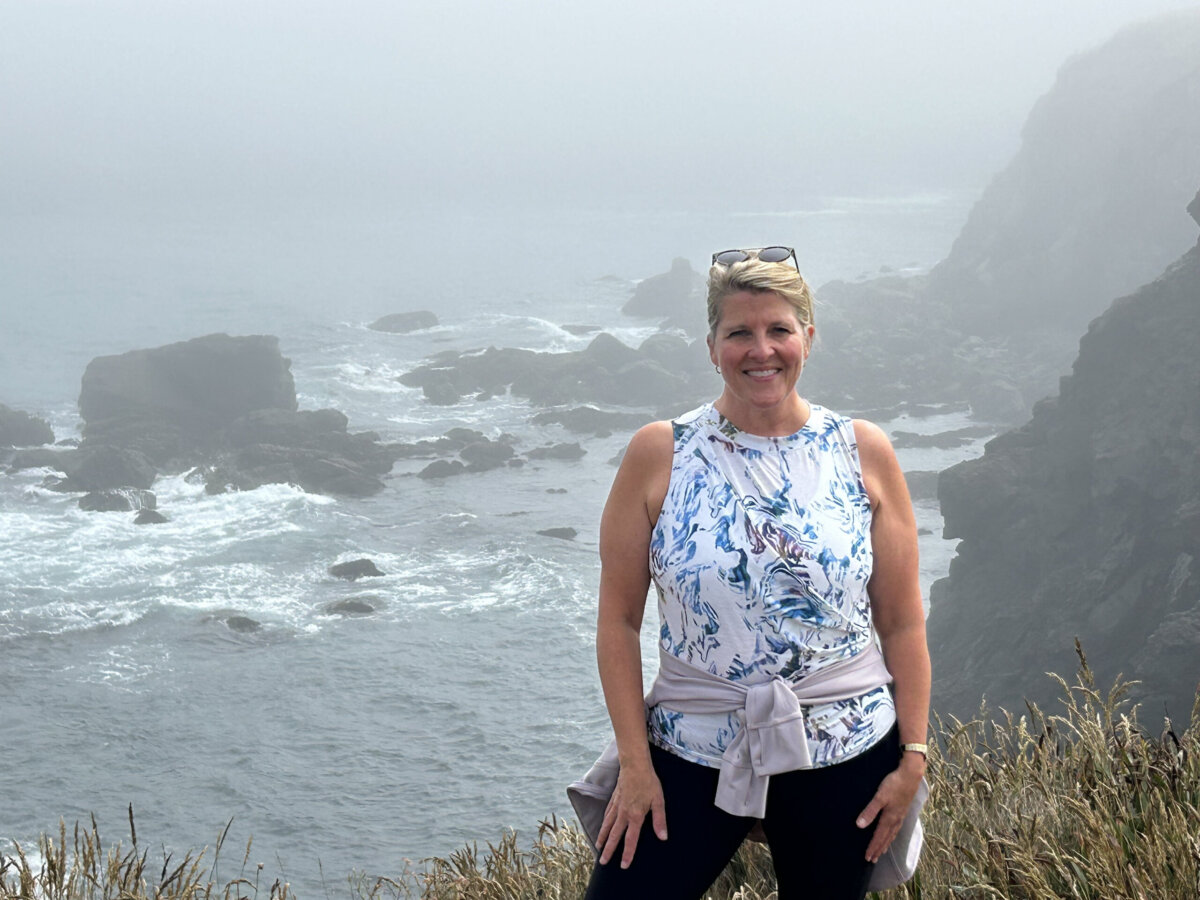 Woman stands smiling by a foggy, rocky ocean coastline, wearing a sleeveless top, leggings, and a sweater tied at her waist.