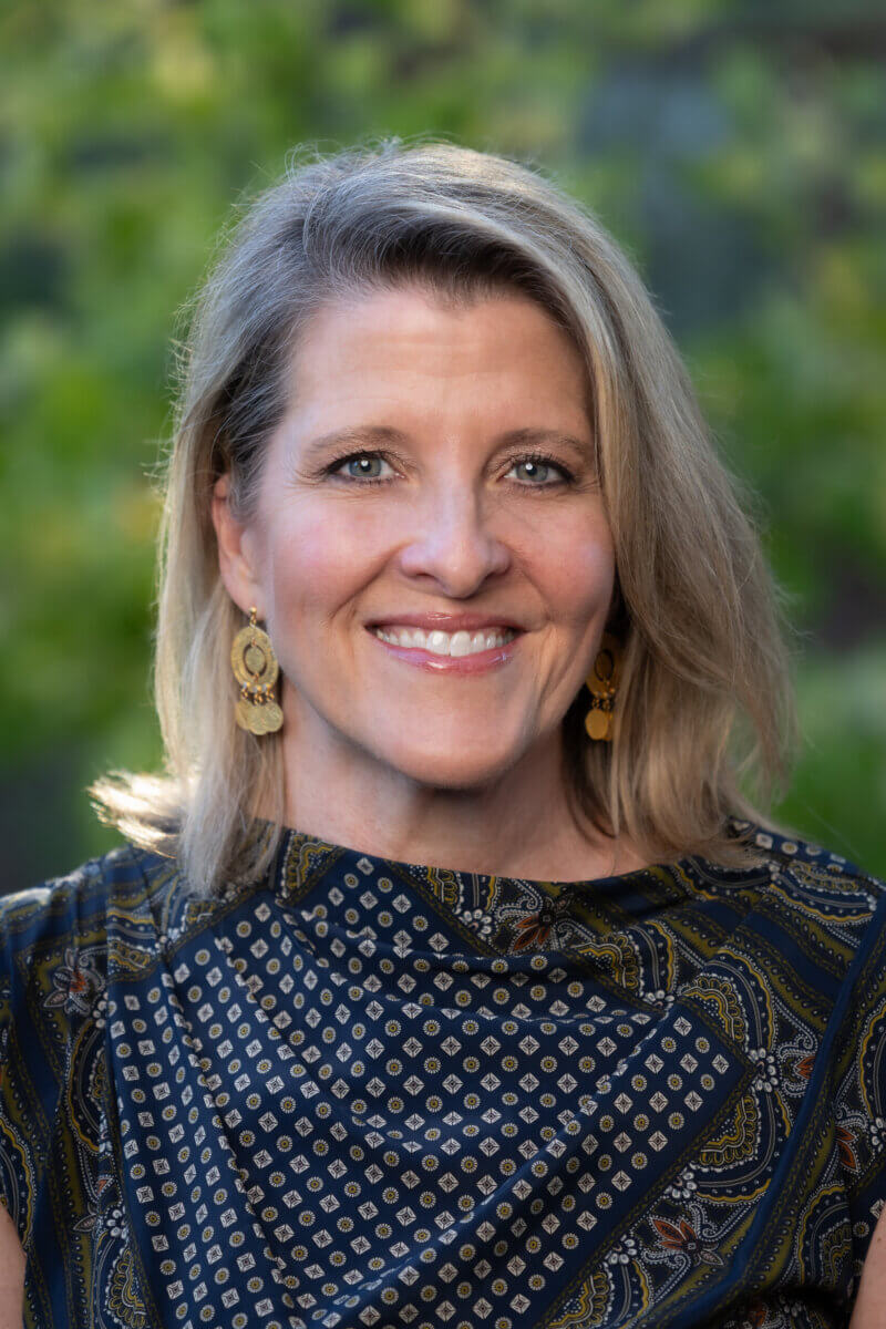 A woman with shoulder-length blonde hair, wearing gold earrings and a patterned top, smiles at the camera.