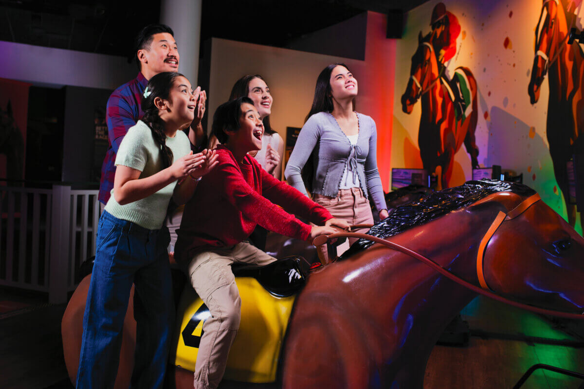 Five people enthusiastically participate in an indoor horse racing arcade game, with one riding a mechanical horse and others cheering. Brightly colored lights and horse murals are visible in the background, making this one of those Southern attractions that’s truly worth the drive.
