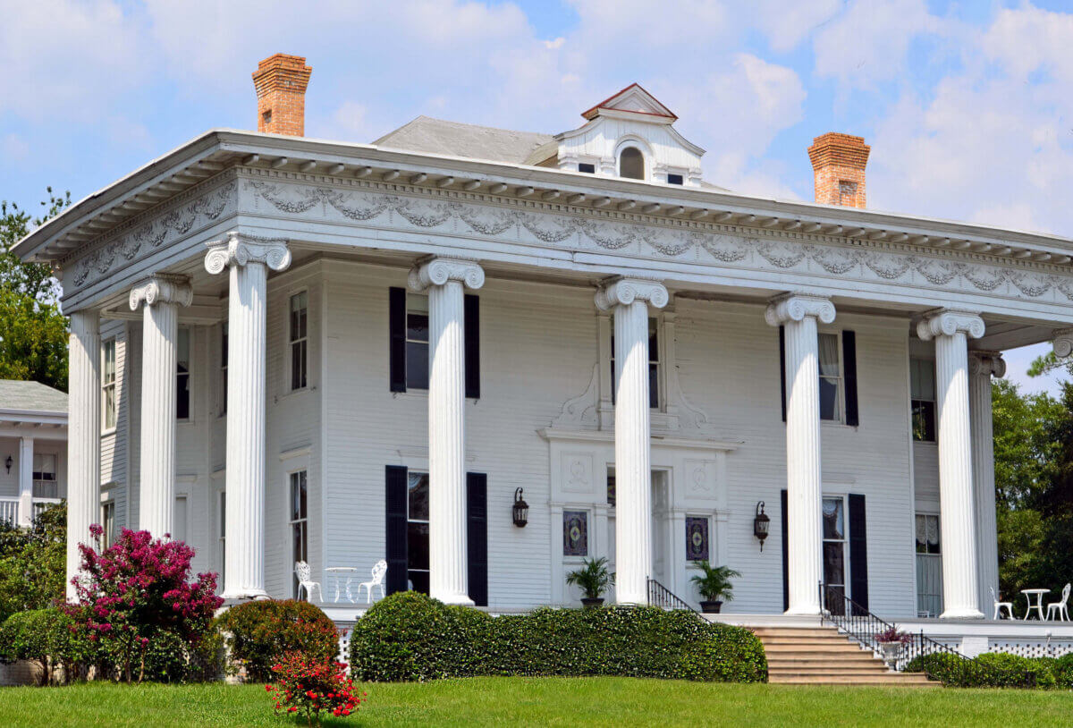 Large white mansion with tall columns, black shutters, and two brick chimneys, surrounded by manicured bushes and flowering shrubs on a sunny day, exuding classic Southern charm with stylish add-ons that enhance its timeless elegance.