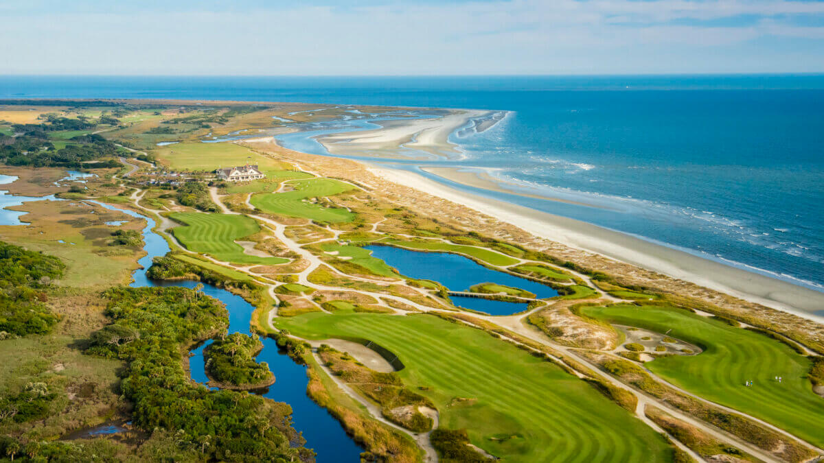 Aerial view of a coastal golf course with green fairways, sand bunkers, ponds, and the ocean shoreline under a partly cloudy sky, exuding Southern Charm throughout its scenic landscape.