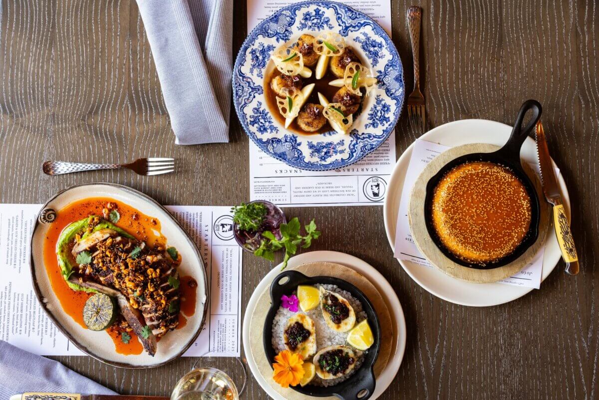 The Best Culinary Hot Spots in the South - 5 Four plated gourmet dishes—including bread, meatballs with sauce, oysters with lemon, and a steak with garnish—are beautifully arranged on a wooden table set with menus and a cloth napkin, capturing the vibrant flavors of the South for an unforgettable experience at one of its best culinary hot spots.